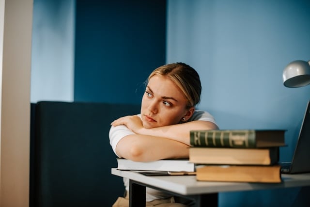 NAD Boosters: What Works and What Doesn't 7 A young woman rests her head on her arms while sitting at a desk with stacked books.