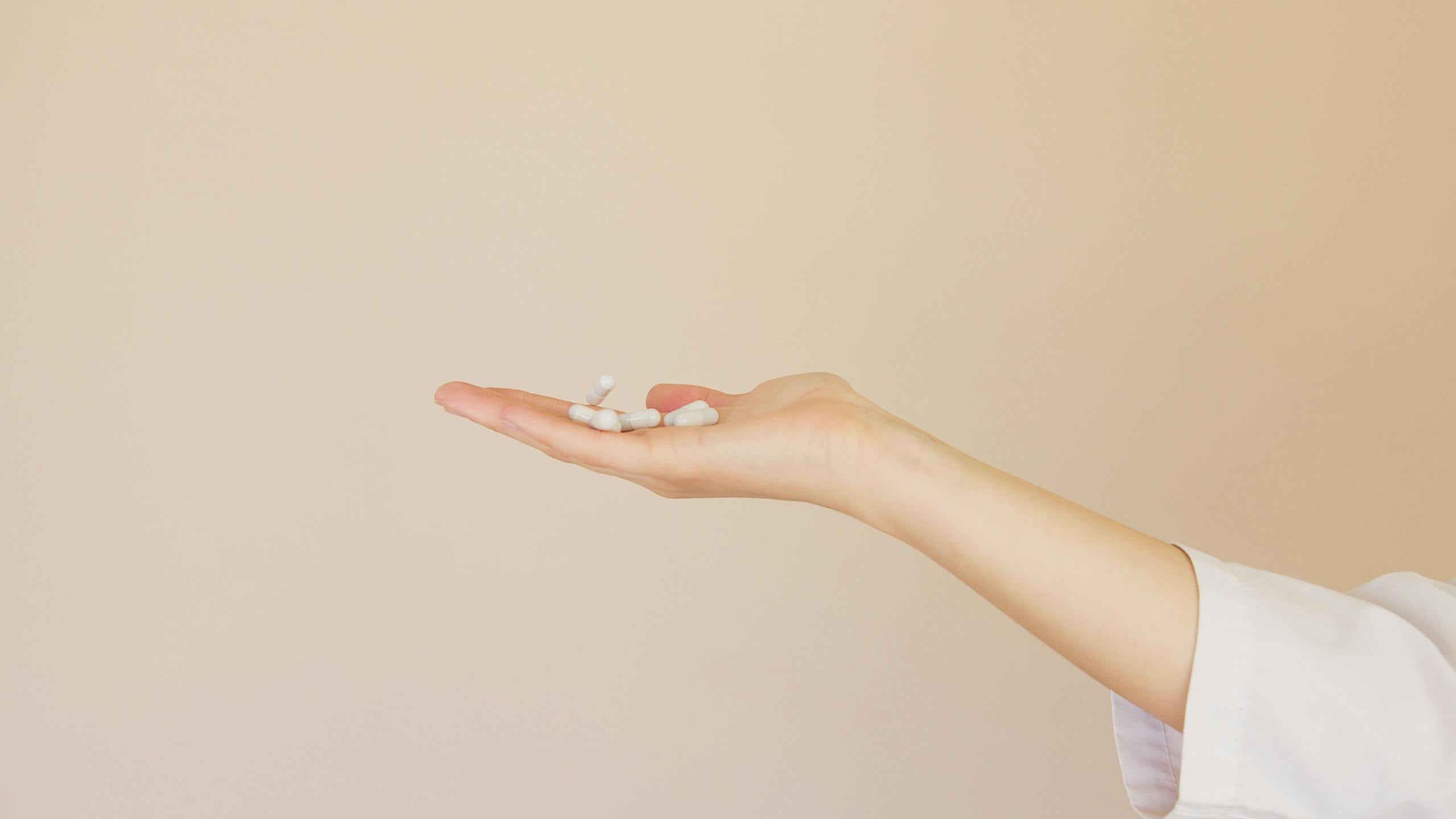NMN with TMG: Benefits, Dosage and Risks 4 A female hand in a lab coat holds white capsules against a neutral background.