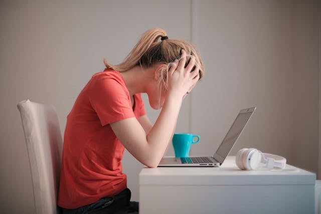 NMN for Brain Fog: Exploring its Role in Cognitive Function 8 A woman in a coral t-shirt sitting at a desk with their head in their hands, appearing stressed or frustrated