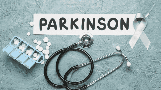 Flat lay image featuring the word "Parkinson" with a white awareness ribbon, pills, a pill organizer, and a stethoscope, symbolizing Parkinson’s disease awareness and healthcare.
