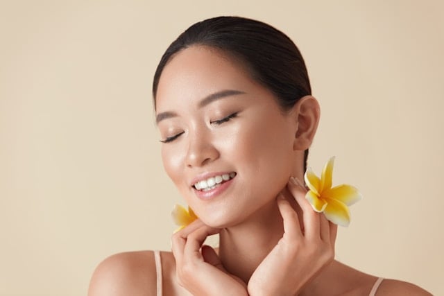 What to Expect Before and After Taking NMN Supplements 7 A smiling Asian woman with closed eyes touches her neck with yellow flowers by her face.