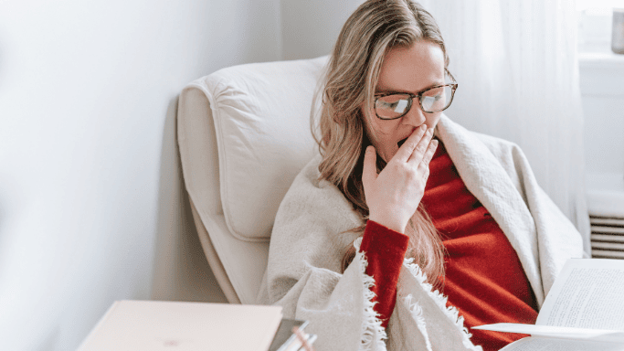 8 Ways to Boost Your Glutathione Levels Naturally 7 An image showing tired woman yawning while reading, wrapped in a blanket—illustrating fatigue, a common sign of low glutathione levels.