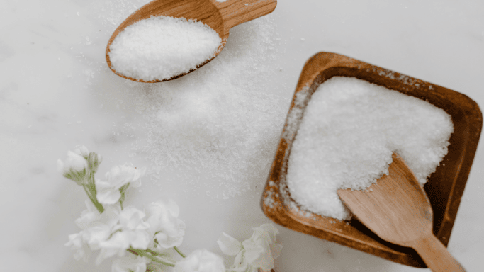 An image showing a wooden spoon and bowl filled with Epsom salt on a marble surface, with white flowers placed nearby.