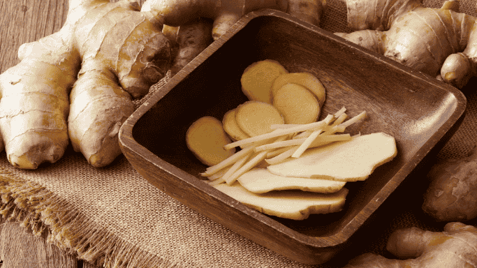 An image showing a fresh ginger root with sliced and julienned ginger pieces in a wooden bowl.