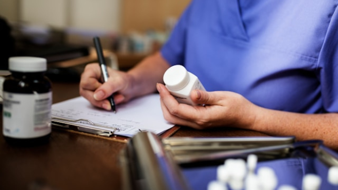 Niacin (B3): Side Effects, Reactions, Overdose Risks Explained 5 An image showing a healthcare professional in blue scrubs holding a pill bottle and writing on a clipboard, with medicine bottles on the table.