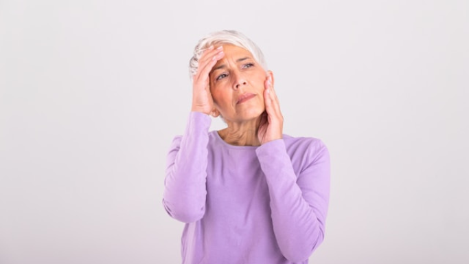 Niacin (B3): Side Effects, Reactions, Overdose Risks Explained 8 An image showing an older woman in a light purple shirt holding her head and face with a pained expression, possibly experiencing a headache or discomfort.