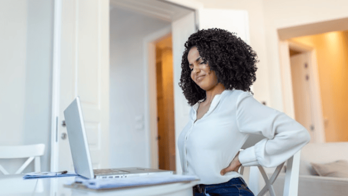 Is Niacin Good for Your Heart? New Studies Show Hidden Risks 9 A woman with curly hair in a white shirt holds her lower back while sitting at a home office desk.