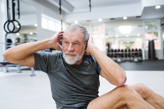 An older man with gray hair and beard does sit-up exercises in a modern gym.