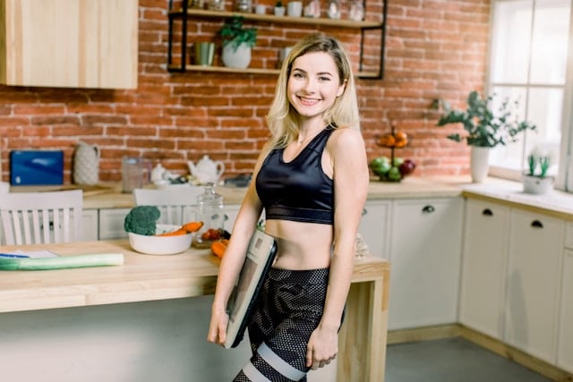 A smiling blonde woman in athletic wear stands in a bright kitchen with vegetables.