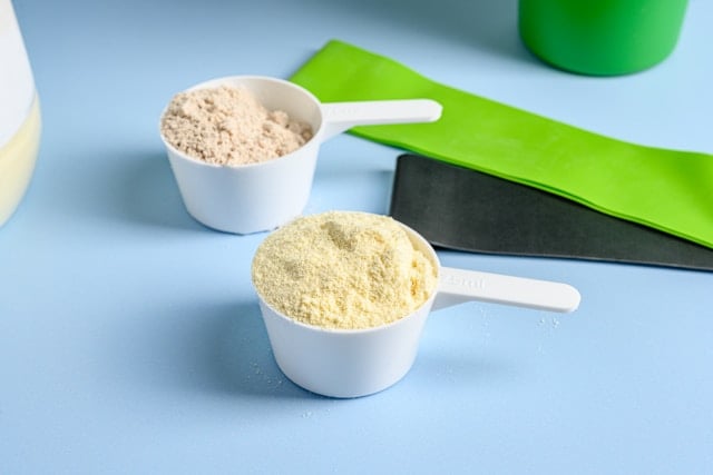 Creatine vs Beta-Alanine: Which is Best for Performance? 4 Two white scoops containing different colored protein powders sit on a blue surface near green items.