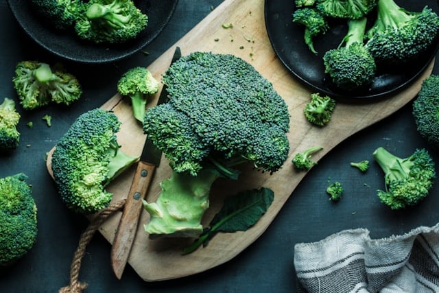 8 Ways to Boost Your Glutathione Levels Naturally 6 Fresh broccoli florets are arranged on a wooden cutting board with black bowls and kitchen utensils nearby.