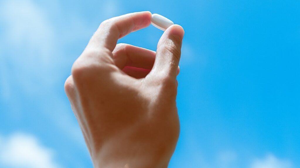 Urolithin A: Benefits, Dosage and Risks You Should Know 6 Hand holding small white supplement capsule between thumb and index finger against bright blue sky with white clouds.