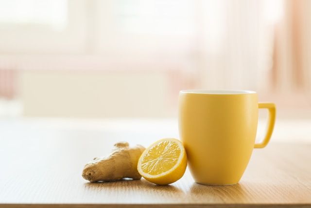 A yellow mug sits beside fresh ginger root and a lemon half on wood.