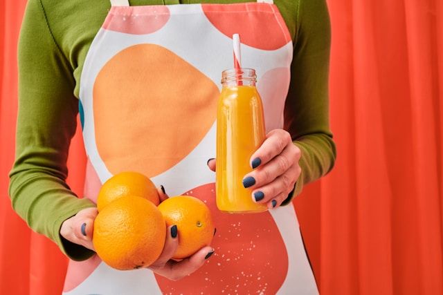 A woman in a green sweater holds fresh oranges and orange juice in a glass bottle.