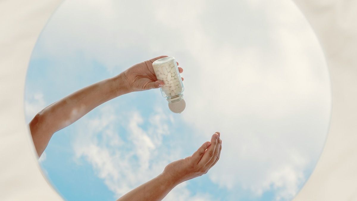 Hand holding small glass vial dispensing white capsules or liquid drops into open palm against blue sky with clouds.