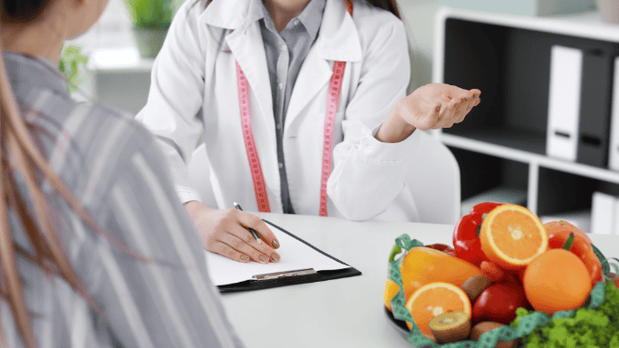 A healthcare professional consults with a patient about diet and health. A bowl of vitamin C-rich fruits sits prominently on the desk, symbolizing the role of nutrition in weight management.