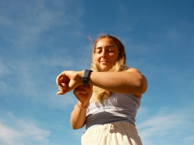 A blonde woman in white clothing checks her smartwatch against a blue cloudy sky.
