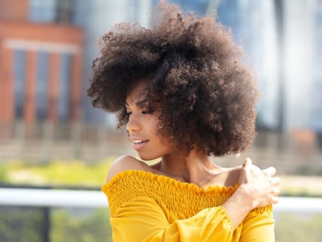 5 Antioxidant Supplements for Oxidative Stress Relief 8 Young woman with voluminous natural curly hair in bright yellow off-shoulder top poses outdoors against blurred urban backdrop.