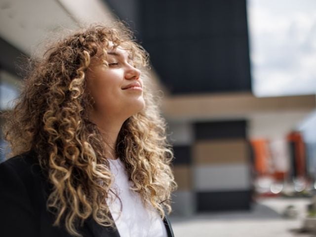 A woman with voluminous curly hair tilts her head back with eyes closed, appearing peaceful in bright natural lighting.Retry