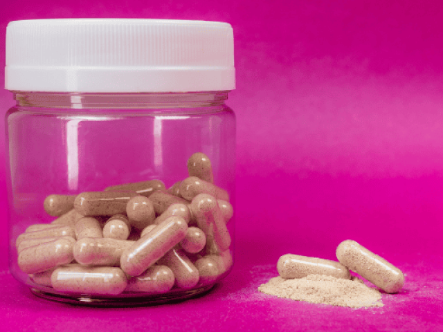 Clear jar of supplements beside capsules and powder on a pink background.