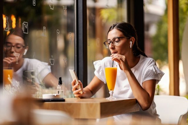 Woman with glasses in white shirt sits at cafe table sipping orange juice.