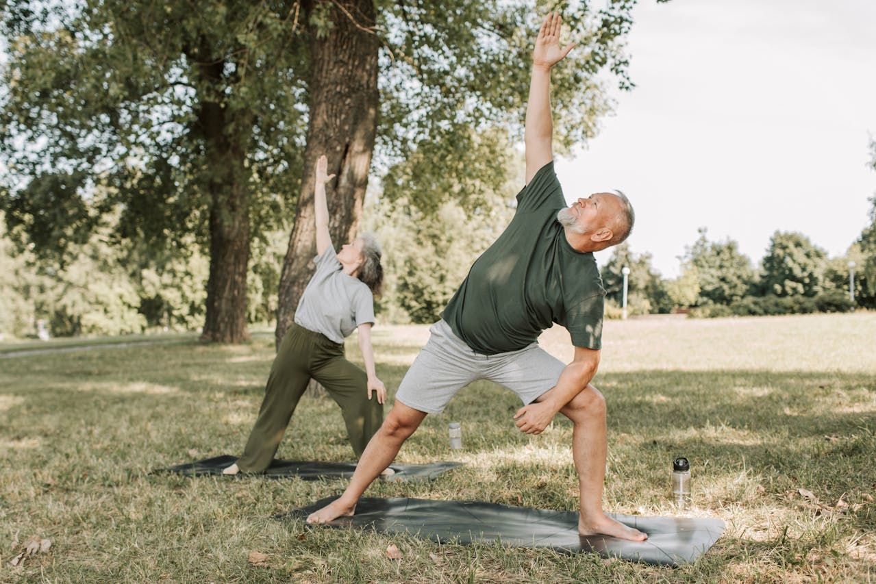Two older adults practicing yoga poses on mats in a grassy park with trees in the background
