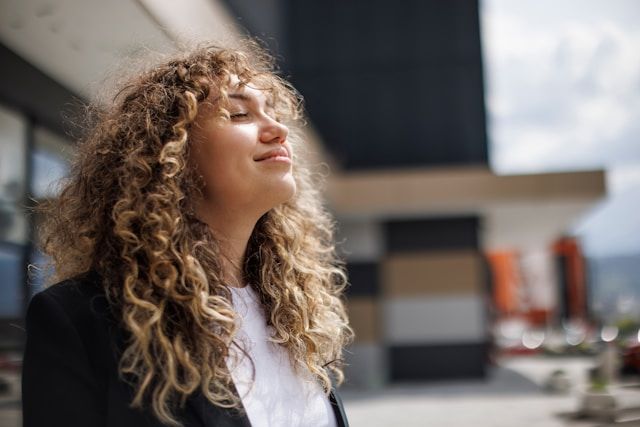 A woman with voluminous curly hair tilts her head back with eyes closed, appearing peaceful in bright natural lighting.Retry