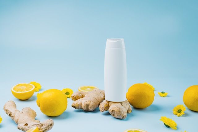 A white bottle sits surrounded by fresh lemons, ginger root, and yellow flowers on blue background.