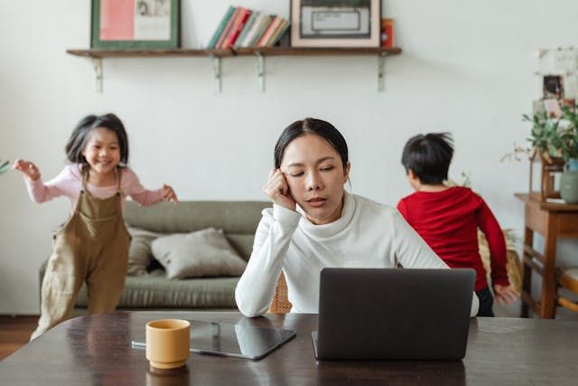 A woman with daytime fatigue at her laptop while her kids play in the background