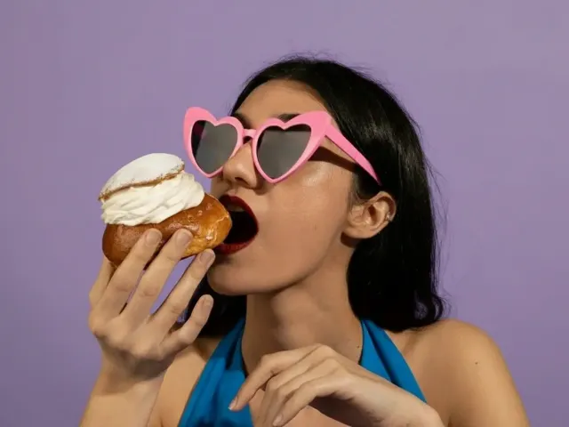 Woman wearing pink heart-shaped sunglasses takes bite of cream-filled pastry against vibrant purple background in playful portrait.