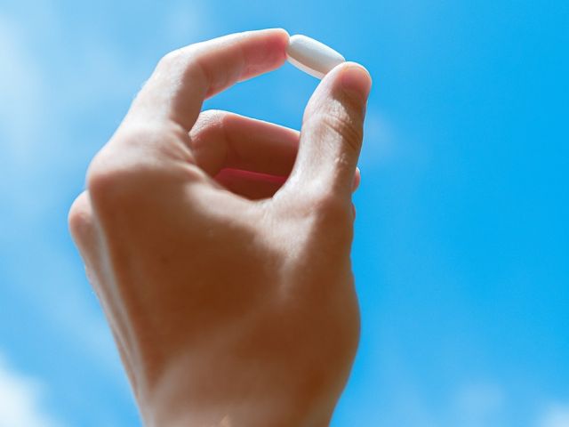 Hand holding small white supplement capsule between thumb and index finger against bright blue sky with white clouds.
