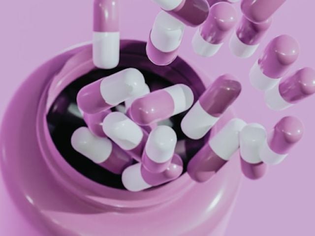 Pink and white capsules spill from a dark circular container onto a matching pink surface.