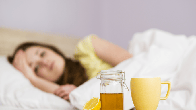 An image showing a sick woman lying in bed with lemon tea on bedside table for cold relief.