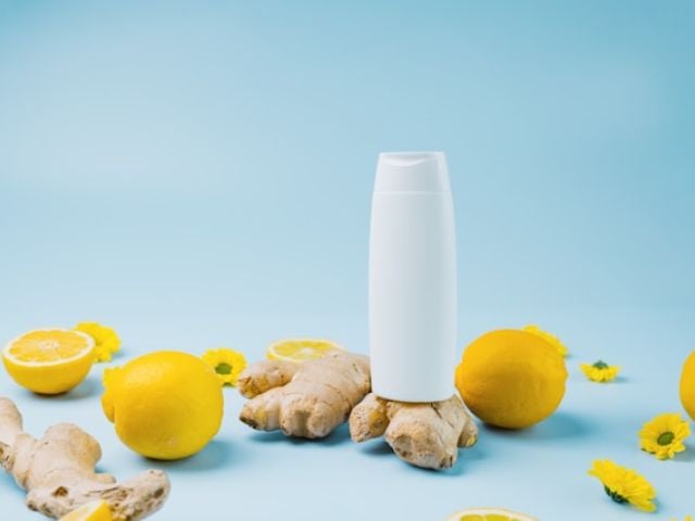 A white bottle sits surrounded by fresh lemons, ginger root, and yellow flowers on blue background.