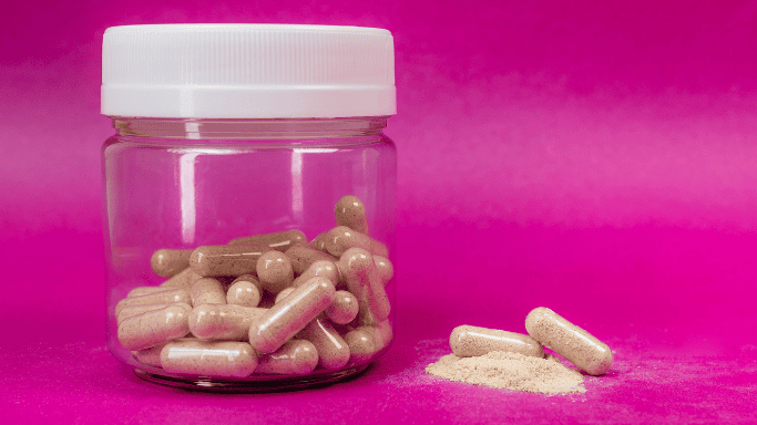 Clear jar of supplements beside capsules and powder on a pink background.