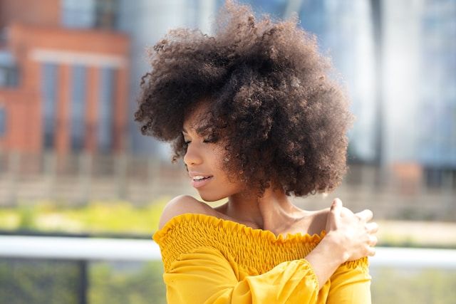 Young woman with voluminous natural curly hair in bright yellow off-shoulder top poses outdoors against blurred urban backdrop.