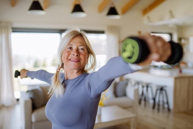 A healthy older woman in a gray top performs arm exercises with dumbbells at home.