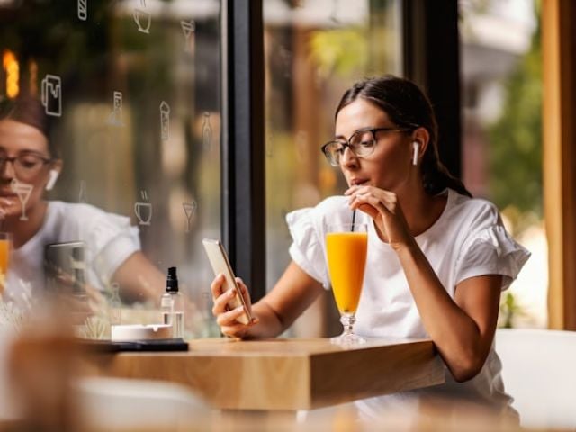 Woman with glasses in white shirt sits at cafe table sipping orange juice.