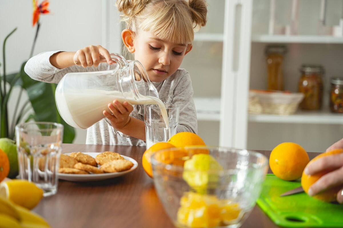 A child pours a glass of milk while her mother cuts oranges in the background.