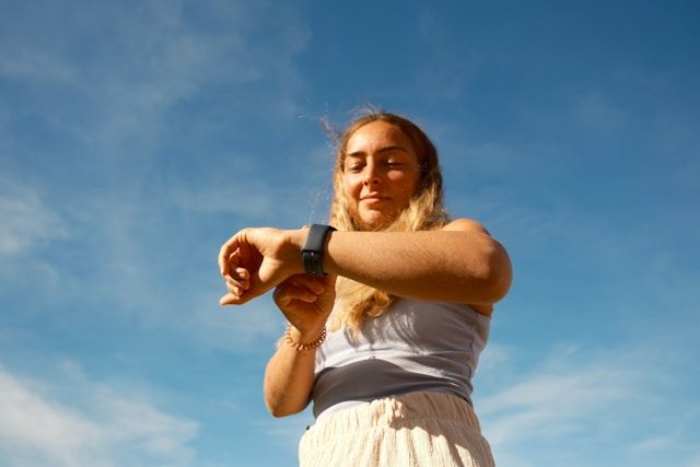 A blonde woman in white clothing checks her smartwatch against a blue cloudy sky.