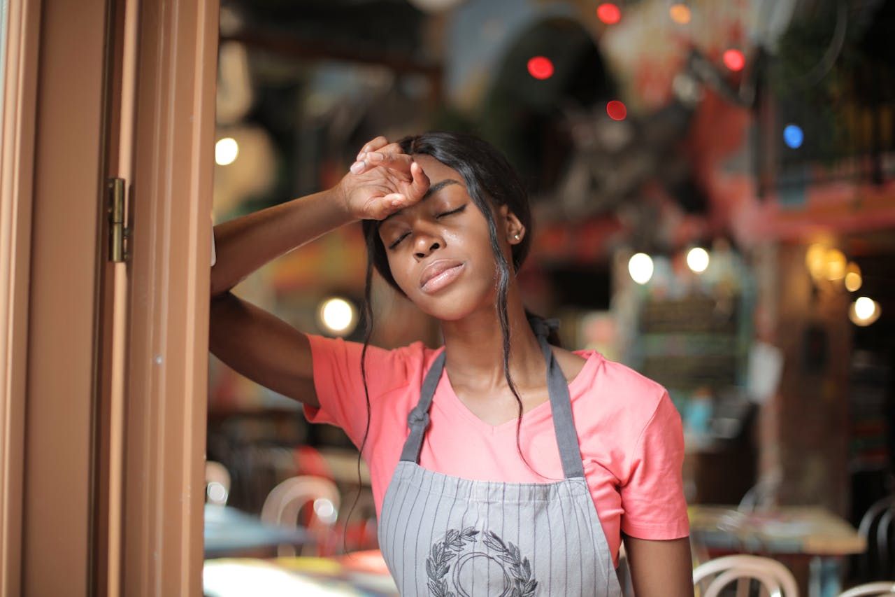 Woman with brain fog wearing a coral pink shirt and apron stands with eyes closed and hand on forehead, appearing tired or stressed