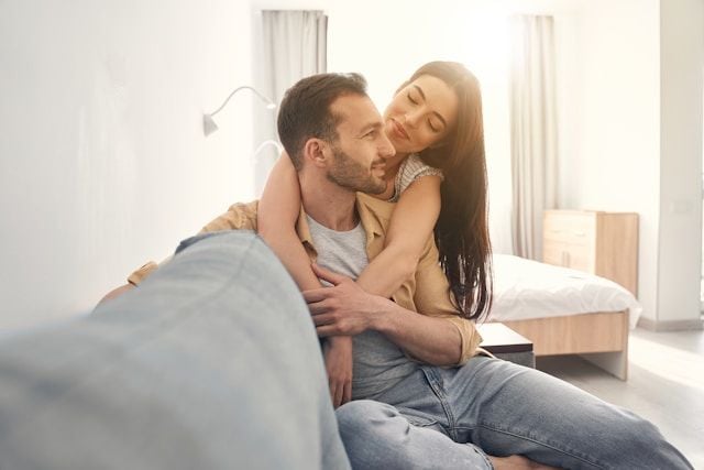 A couple embraces lovingly while sitting together on a couch in bright sunlight.