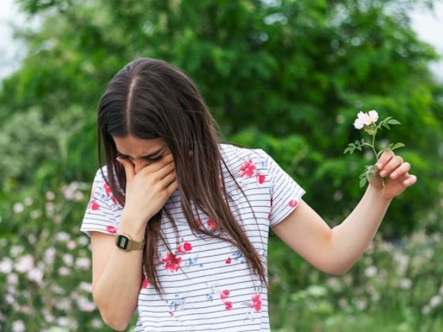 A young woman holds a small flower while covering her face, appearing to have an allergic reaction outdoors.