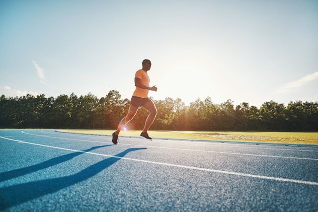 A runner sprints on a blue athletic track during golden hour with trees and sky visible.