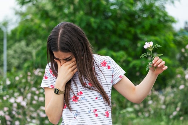 A young woman holds a small flower while covering her face, appearing to have an allergic reaction outdoors.