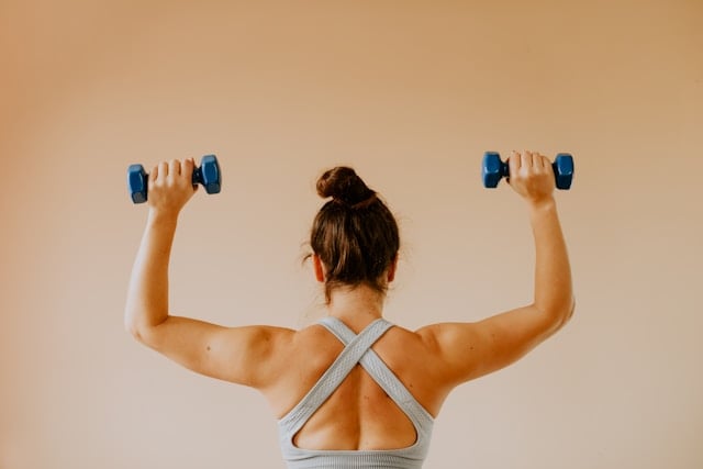 NAD Boosters: What Works and What Doesn't 7 A woman with hair in a bun lifts blue dumbbells overhead from behind view.