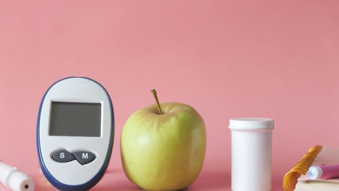 A blood sugar monitor, apple and pill container on a pink background