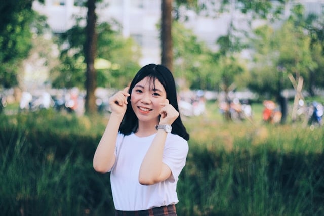 A smiling young Asian woman in a white shirt poses with hands near her face in a park setting.