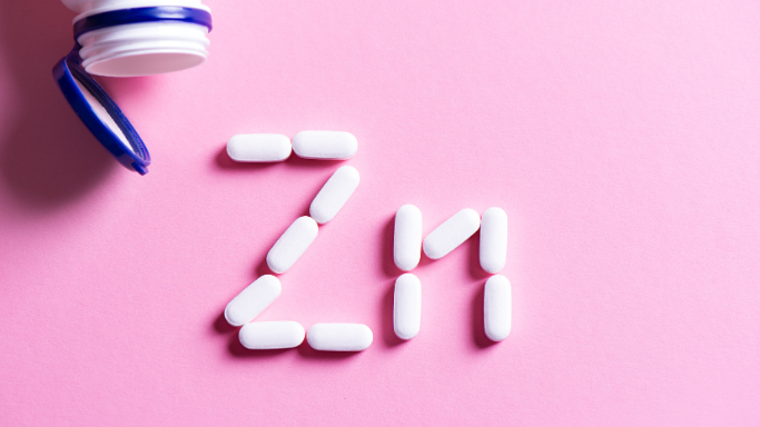 An image showing white pills spelling "Zn" on a pink background, with an open pill bottle nearby, representing zinc supplements.