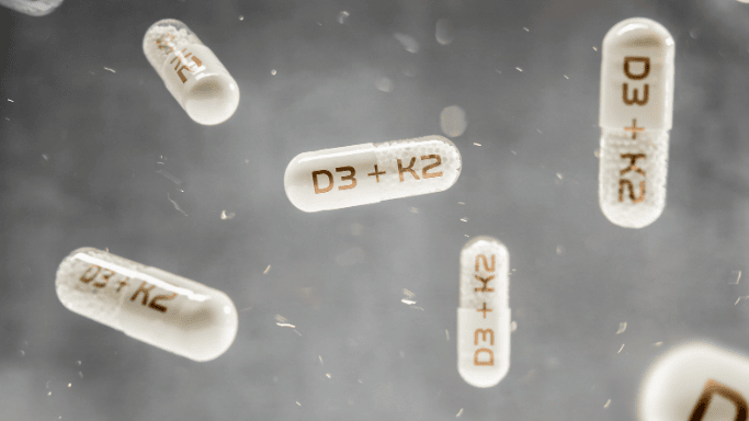 An image showing several white capsules floating against a blurred gray background. Each capsule is labeled "D3 + K2".